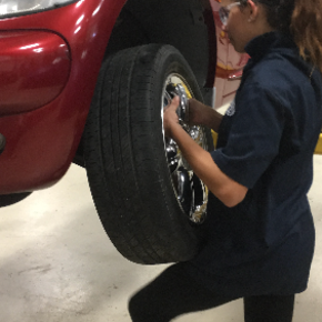 A female student changing the tire on a car that is up on a lift. 